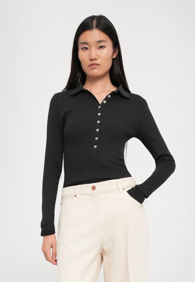 O femeie cu păr lung, drept și negru poartă un top negru cu mânecă lungă, ajustat, și pantaloni albi of-white cu talie înaltă, cu mâna în buzunar.