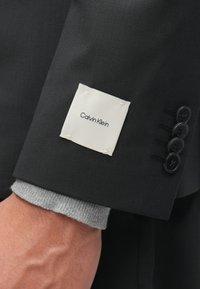 Μαύρο σακάκι με λευκή ετικέτα ραμμένη στο μανίκι που λέει "Calvin Klein." Έχει τέσσερα μαύρα κουμπιά και γκρι πλέξη στο μανίκι.