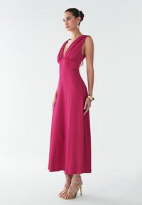 Fuchsia, sleeveless maxi dress with a deep V-neck and gathered bodice, featuring a fitted waist and flowing skirt, paired with strappy heels.