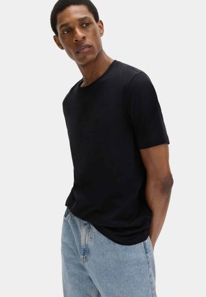 Mężczyzna ubrany w prostą czarną koszulkę z krótkim rękawem i jasnoniebieskie dżinsy, stojący z rękami za plecami, lekko patrzący na bok.