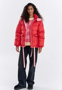 Czerwona kurtka puchowa z różowymi akcentami, z kapturem, elastycznymi mankietami i troczkami. Noszona na różowym swetrze w paski i ciemnych spodniach.