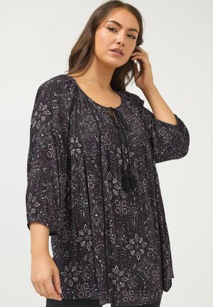 Kobieta ubrana w luźną, czarną bluzkę z długim rękawem, ozdobioną białym i fioletowym wzorem paisley oraz troczkami z frędzlami przy dekolcie.