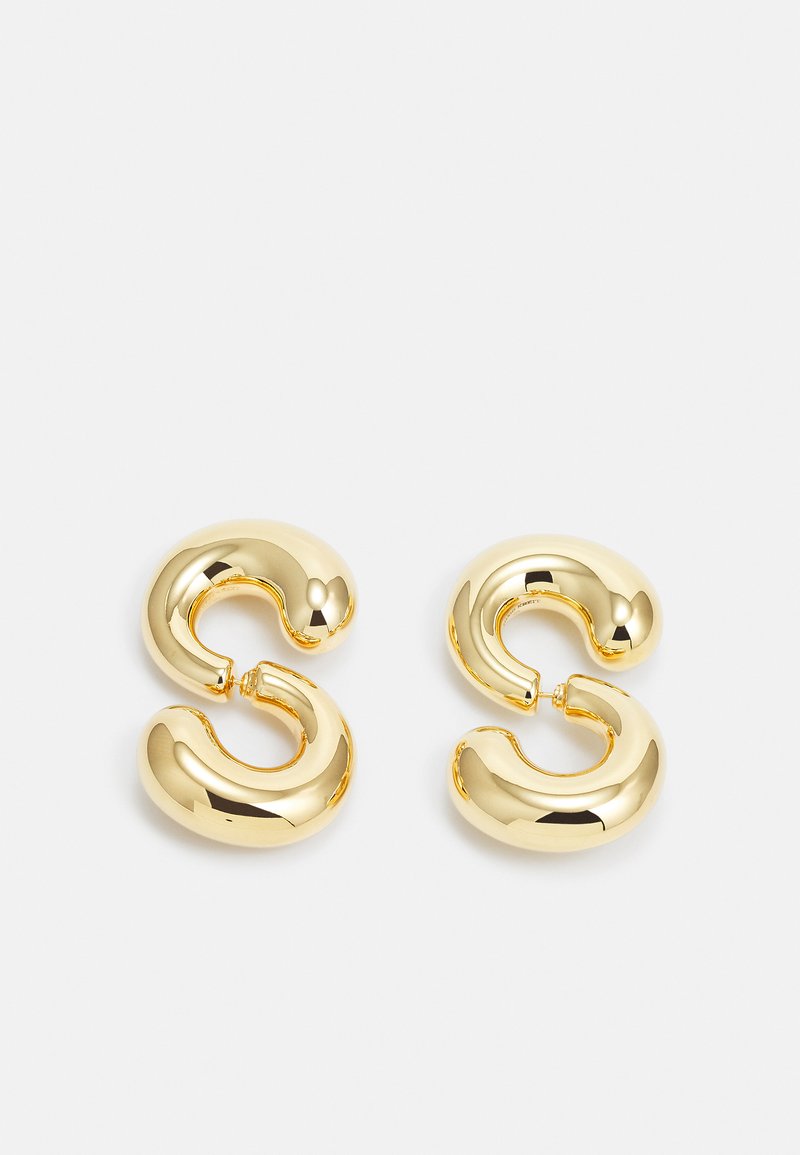 Brincos em argola de tom dourado com uma superfície lisa e brilhante; apresentam um design curvado e abstrato que se assemelha a uma letra "S" estilizada.