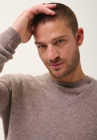 Světle šedý žebrovaný pletený svetr s výstřihem do V. Model jemně sahá na hlavu a předvádí měkkou texturu a uvolněný střih.