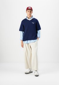 Pull en tricot bleu marine à manches courtes sur une chemise à rayures bleues, associé à un pantalon large crème et des baskets noir et blanc, complété par une casquette rouge.