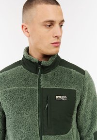 Joven con el cabello corto que lleva una chaqueta de forro polar verde con cremallera y un bolsillo en el pecho etiquetado "PROTESTA," mirando hacia un lado.