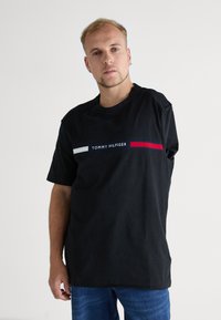 Crna pamučna majica s okruglim izrezom, s grafikom bijelih i crvenih pruga s tekstom "TOMMY HILFIGER" u sredini. Ležerni kroj.