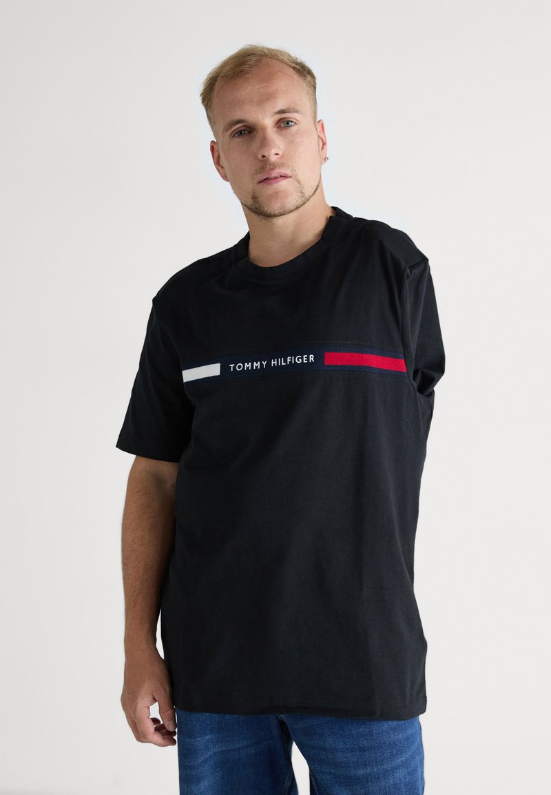 Crna pamučna majica s okruglim izrezom, s grafikom bijelih i crvenih pruga s tekstom "TOMMY HILFIGER" u sredini. Ležerni kroj.