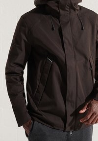 Brązowa wodoodporna kurtka z kapturem, z zamkiem błyskawicznym z przodu i bocznymi kieszeniami. Charakteryzuje się gładką fakturą i regulowanymi sznurkami.