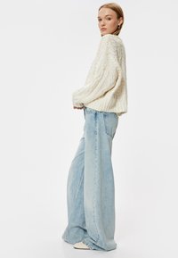 Cremfarbener Kabelstrickpullover, lässige Passform; hellblaue weit geschnittene Jeans. Weiche Textur, voluminöses Design, leichte Abnutzung an den Stoffkanten.