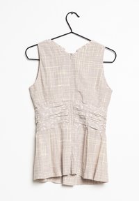 Top senza maniche di un leggero beige con un motivo a griglia testurizzato, cerniera sul retro e dettagli arricciati sui lati, appeso a un gancio nero.