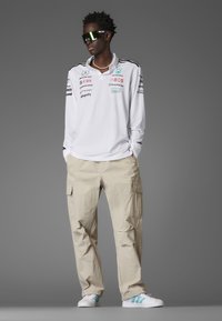 White long-sleeve shirt with black and colored logos, beige cargo pants, and white shoes with teal stripes. Reflective sunglasses complete the look.