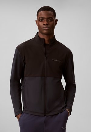 Mand iført en sort Calvin Klein jakke med lynlås, med hænderne i lommerne på marineblå bukser mod en ensfarvet grå baggrund.