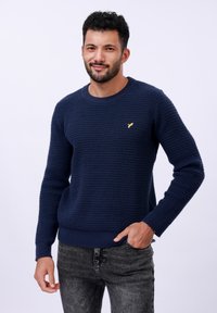 Pull en maille nid d'abeille bleu marine avec un col rond, des manches longues et un petit logo jaune sur la poitrine. Assorti à un jean gris foncé.