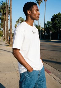 Joven con camiseta blanca y vaqueros azules en una acera soleada bordeada de palmeras, con las manos en los bolsillos, mirando hacia la derecha.