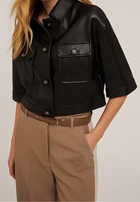 Giacca in pelle nera corta con maniche corte, due tasche sul petto e bottoni metallici; abbinata a pantaloni beige a vita alta e una cintura.