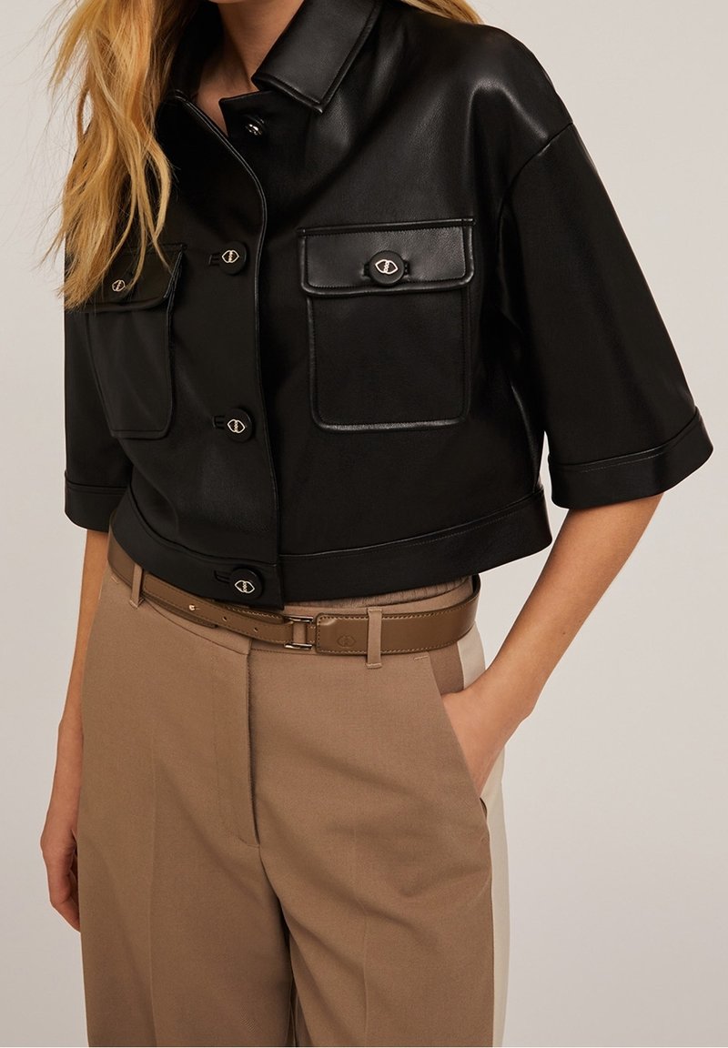 Giacca in pelle nera corta con maniche corte, due tasche sul petto e bottoni metallici; abbinata a pantaloni beige a vita alta e una cintura.