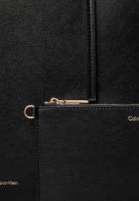 Sac en cuir noir texturé avec fermeture éclair dorée et logo Calvin Klein doré, doté d'une attache de bracelet amovible.