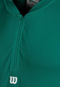 Maglietta sportiva verde con zip, realizzata in tessuto testurizzato, con una zip frontale e un logo bianco sul lato sinistro del petto.