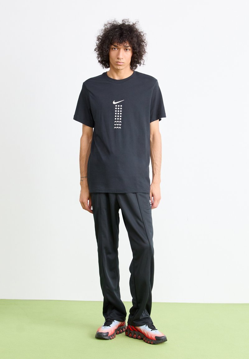 Zwarte katoen T-shirt met wit Nike-logo en patroonafbeelding, gecombineerd met zwarte sportbroeken en meerkleurige hardloopschoenen.