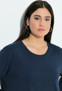 Marineblaues langärmeliges Shirt mit rundem Halsausschnitt und weichem Textil. Das Model trägt silberne Creolen und hat einen neutralen Hintergrund.