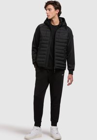 Zwarte gewatteerde vest met capuchon, met gestikte secties en een rits aan de voorkant, gecombineerd met zwarte joggingbroeken en witte sneakers.