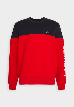 Sudadera con cuello redondo con sección superior negra, sección inferior roja, logotipo de Lacoste en el pecho y texto "LACOSTE" en la manga izquierda en blanco.