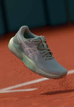Baskets de sport gris clair avec tige en maille, accents roses et semelle texturée, présentées en l'air au-dessus d'une surface en argile rouge.