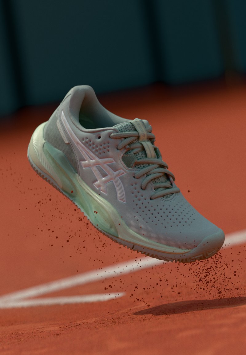 Baskets de sport gris clair avec tige en maille, accents roses et semelle texturée, présentées en l'air au-dessus d'une surface en argile rouge.