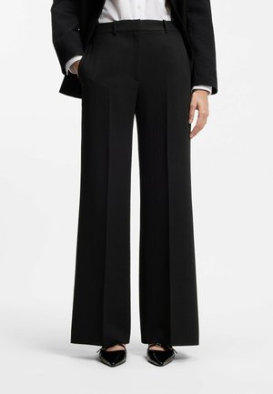 Kvinde iført sorte brede, skræddersyede bukser, sort blazer, hvid skjorte og sorte spidse sko med ankelremme.