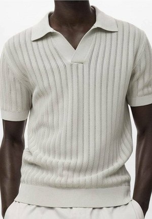 Dark-skinned person wearing a short-sleeve beige knit polo shirt with vertical zigzag patterns and light-colored pants.