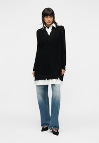 Μαύρη πλεκτή τουνίκ με V-λαιμόκοψη, φορεμένη πάνω από λευκό πουκάμισο, σε συνδυασμό με ανοιχτό μπλε τζιν φαρδιά στα μπατζάκια και μαύρα μυτερά παπούτσια.