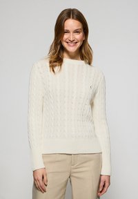 Cremfarbener Strickpullover mit Kabelmuster und geripptem Saum sowie Bündchen. Weiche Textur, runder Halsausschnitt, mit kleinem besticktem Logo auf der Brust.