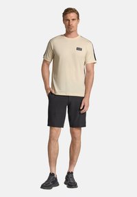 Beige t-shirt med rund halsringning och korta ärmar, prydd med en svart logotyplapp, tillsammans med svarta shorts och svarta träningsskor.