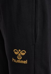 Schwarze Jogginghose aus strukturiertem Stoff. Mit einem goldfarbenen Logo mit einem Bienen-Design und dem Text "hummel" in der Nähe der linken Seitentasche.