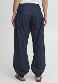 Pantalones ligeros azul marino con cintura fruncida y piernas anchas y cónicas. Presentan puños elásticos y una tela lisa y texturizada.