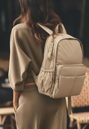Una persona che indossa abbigliamento beige a coste porta uno zaino beige coordinato con multiple tasche e spallacci regolabili.