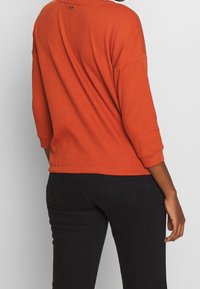 Ribbad, rostorange långärmad tröja med sänkta axlar och rundad nederkant. Bärs med svarta jeans som visar upp en avslappnad passform och textur.