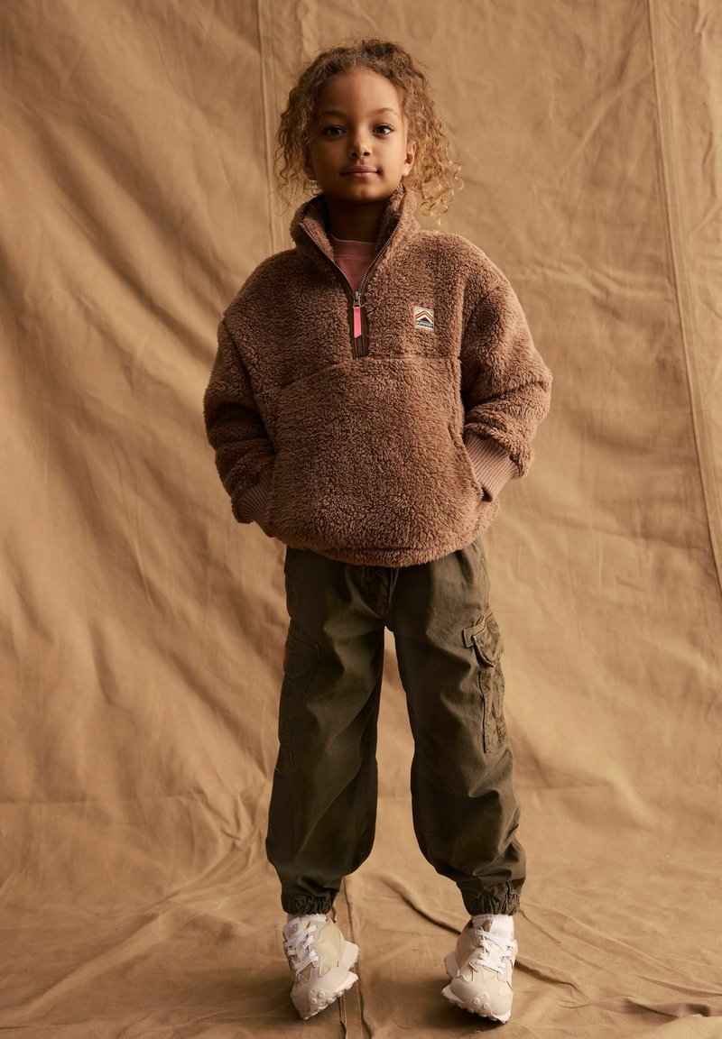 Pullover di pile marrone con colletto alto e zip a metà, abbinato a pantaloni cargo verdi con polsini elasticizzati e tasche, indossato con sneakers bianche.