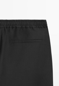Gros plan sur un pantalon noir montrant une ceinture élastique et une seule poche arrière horizontale avec une ouverture biaisée sur fond uni.