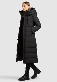 Hosszú fekete puffermellény lehúzható kapucnival, oldalsó zsebekkel és cipzáras elülső résszel. Steppelt kialakítással és tartós, sima textúrával rendelkezik.