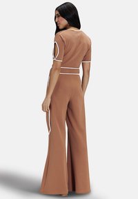 Brown jumpsuit made of stretchy fabric, featuring short sleeves, cut-out details, and white accents. Flared legs enhance the overall design.