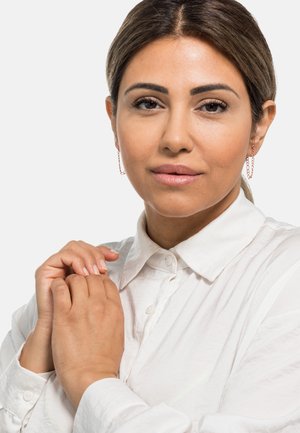 Ohrringe aus Roségold mit einem subtilen Kettendesign, getragen zu einem weißen Hemd mit Kragen. Glatte Hauttextur und natürliches Make-up.