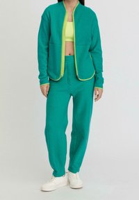 Giacca in pile e pantaloni jogger in turchese brillante con accenti verdi lime a contrasto. La giacca presenta una chiusura a zip e tasche. Le sneakers sono bianche.