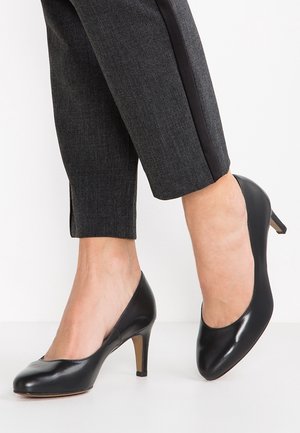 Czarne skórzane szpilki z zaokrąglonym noskiem, o gładkiej fakturze i smukłym, skośnym obcasie. W zestawie z szarymi spodniami.