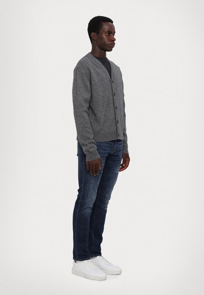 Grau gestrickte Cardigan mit V-Ausschnitt, Knopfverschluss und gerippten Bündchen, kombiniert mit dunkelblauen Jeans und weißen Sneakers.