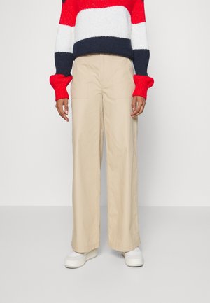 Pantalon cargo beige à jambes larges avec deux poches avant et une coupe droite, associé à un pull en tricot rayé rouge, blanc et bleu marine et des chaussures blanches.
