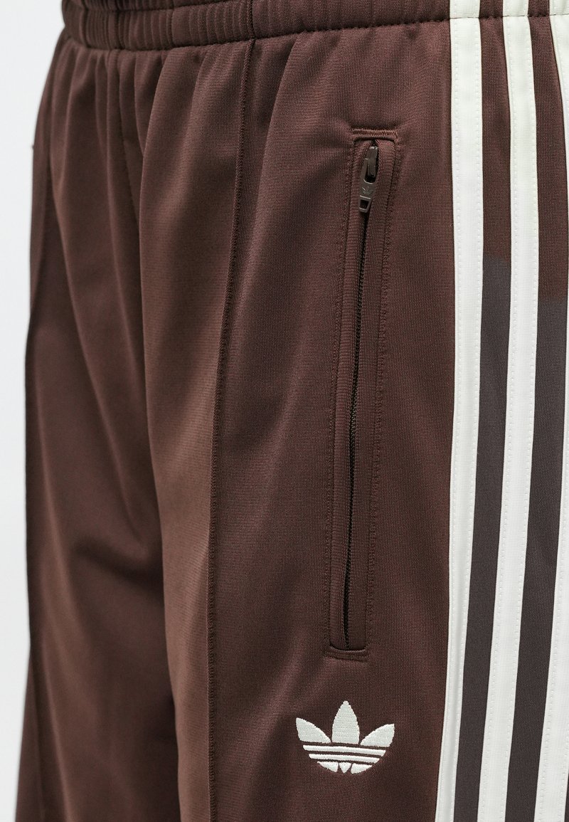 Pantalones deportivos marrones con rayas verticales blancas y grises, un bolsillo con cremallera y un logo bordado en el costado. Fabricados con tela texturizada.