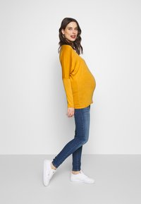 Top mostarda de mangas longas com um corte solto, combinado com jeans skinny azul escuro e ténis brancos. Design simples, textura suave.