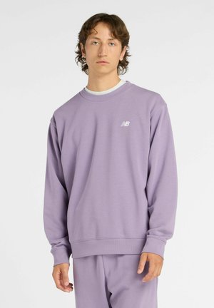 Giovane uomo che indossa una felpa New Balance lilla e pantaloni abbinati, in piedi contro uno sfondo bianco semplice.